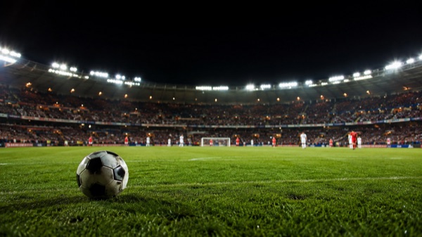 Estadio de fútbol de noche con las tribunas llenas y una pelota sobre el césped en primer plano