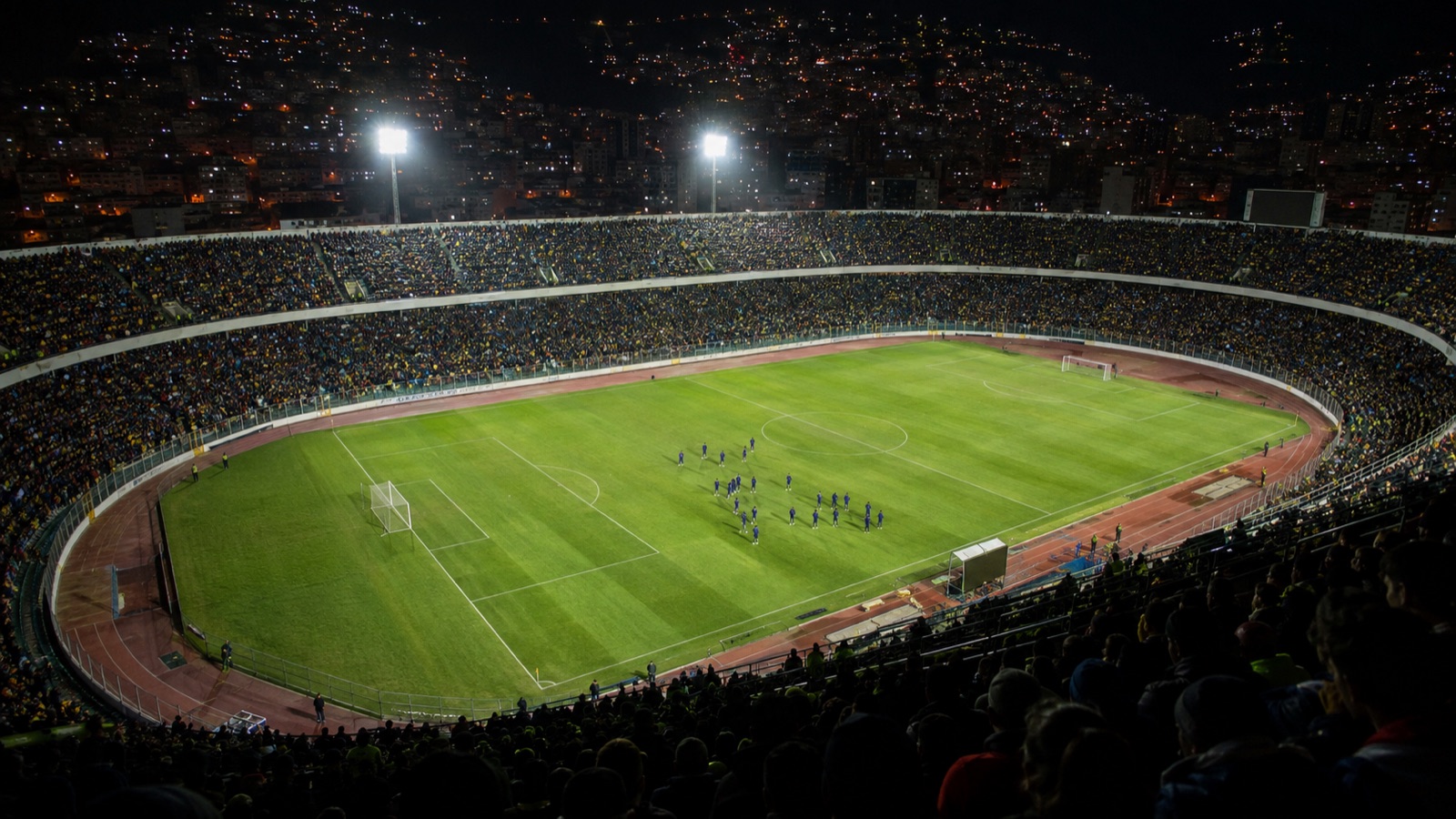 Vista general de un estadio de fútbol de noche, con las tribunas llenas y jugadores en el campo antes de un partido internacional.
