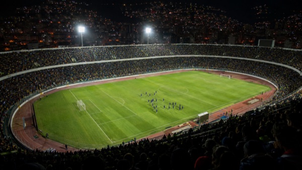 Vista general de un estadio de fútbol de noche, con las tribunas llenas y jugadores en el campo antes de un partido internacional.