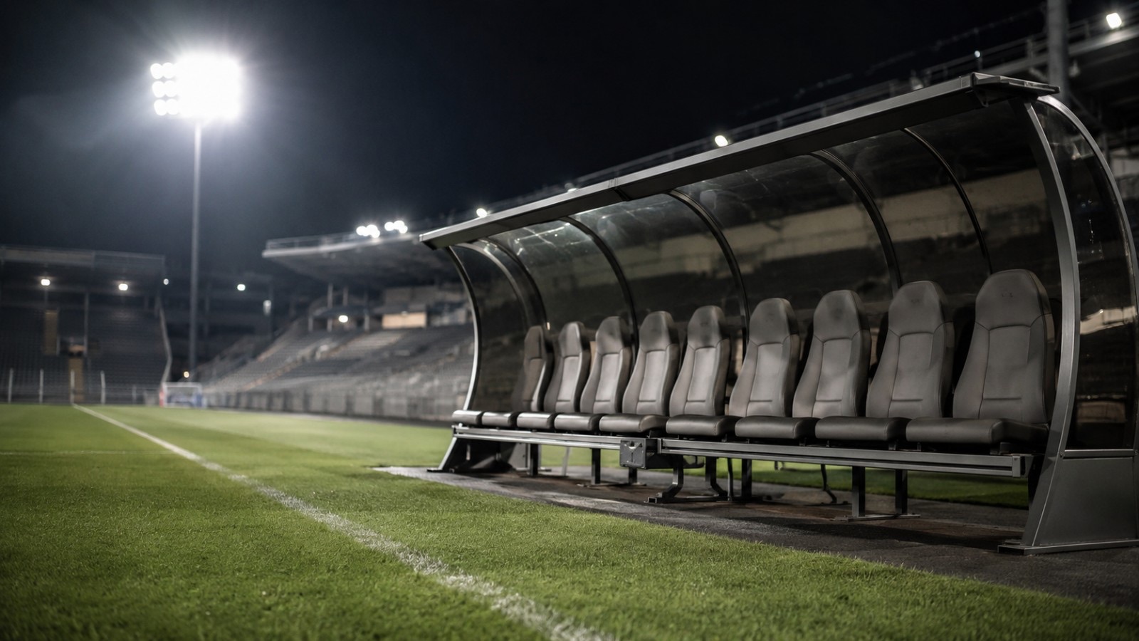 Banco de suplentes vacío al costado de una cancha de fútbol, de noche y con las luces del estadio encendidas
